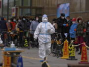 A health worker wearing a protective suit walks by masked residents who wait in line to get their throat swab at a coronavirus testing site following a COVID-19 case was detected in a residential buildings, Wednesday, April 6, 2022, in Beijing.