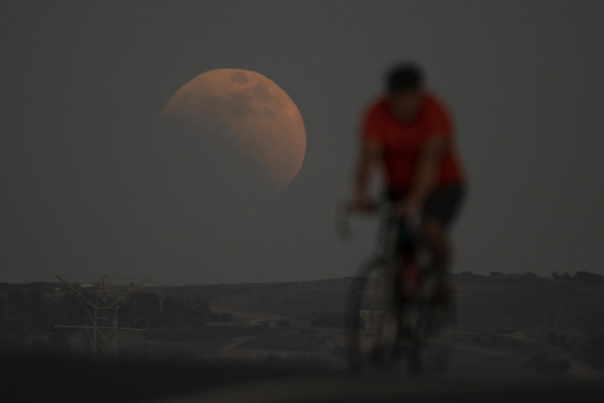 A lunar eclipse is seen behind a cyclist during the first blood moon of the year, in Irwindale, Calif., Sunday, May 15, 2022. (AP Photo/Ringo H.W.