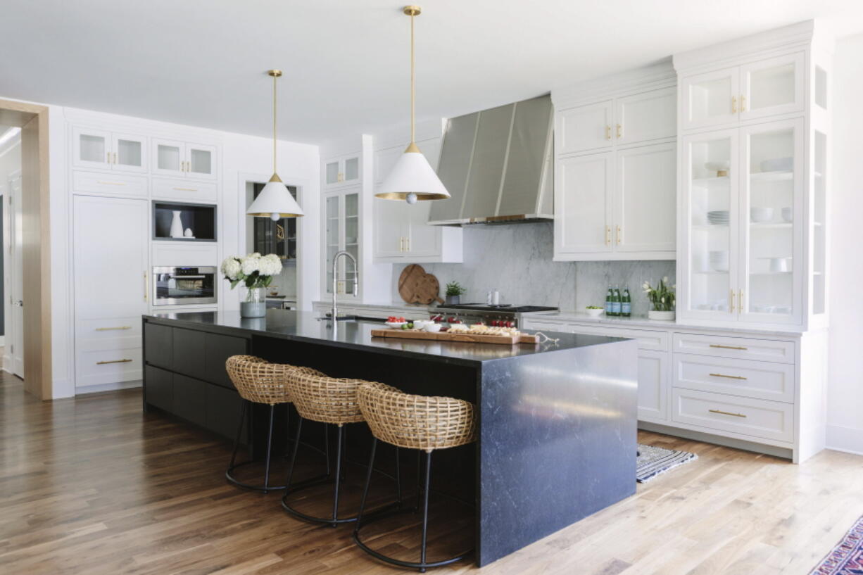 A kitchen design with woven Palecek kitchen stools and a striking midnight blue stone island to balance the extensive Shaker-style white and glass cabinetry. The brass-toned lining of the pendant fixtures from Circa Lighting is echoed in the cabinet hardware.