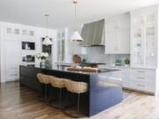 A kitchen design with woven Palecek kitchen stools and a striking midnight blue stone island to balance the extensive Shaker-style white and glass cabinetry. The brass-toned lining of the pendant fixtures from Circa Lighting is echoed in the cabinet hardware.