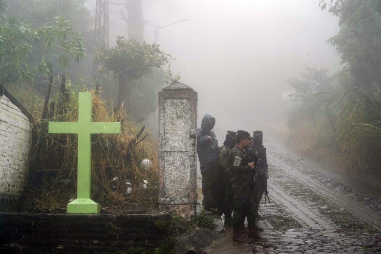 Soldiers stand guard under the rain near a cross that decorates a road in Comasagua, El Salvador, Monday, Oct. 10, 2022. At least five soldiers died on Monday nearby when the wall of a home collapsed as they were sheltering from the rain while deployed to crackdown on gang activity.