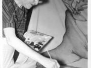 An unknown woman on an unspecified date paints a likeness of the old Hudson's Bay Company flag for an event at the Fort Vancouver site. The visible first letter of the Hudson's Bay Company's Latin motto shows. The phrase "Pro Belle Cutum" literally means "for beautiful skin." Not fully visible are the two stags and four beavers that are part of the Hudson's Bay Company coat of arms and clarify the kinds of skin intended.