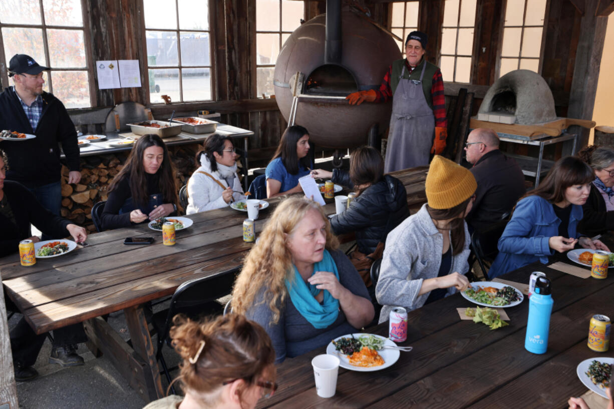 Classmates sit and eat Pasta al Forno, an Italian dish made by Robin Morgan, a PhD student at the Bread Lab, and two types of salad during the Pizza for Producers Training at the WSU Bread Lab in Burlington, Washington, on Oct. 28, 2022. The pasta dish was made in a wood-fired oven converted from a World War II Anti-Submarine Net Buoy seen in the back.