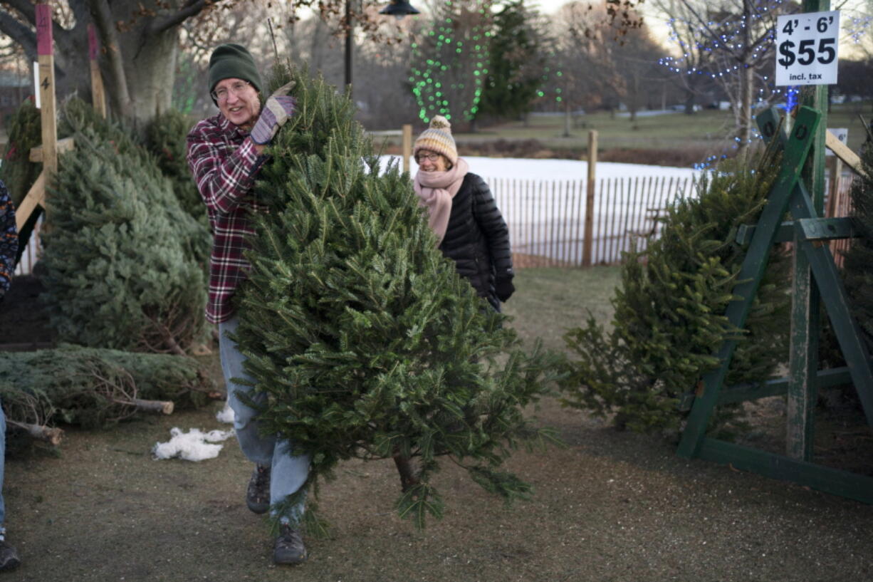 Larry Gurnee carries a $55 Christmas tree he selected with his wife, , Libby Gurnee, background, at a Rotary Club tree sale, Wednesday, Dec. 14, 2022, in South Portland, Maine. Inflation has Americans cutting back on spending in some areas this holiday season but Christmas trees is not one of them, according to the National Christmas Tree Association. (AP Photo/Robert F.