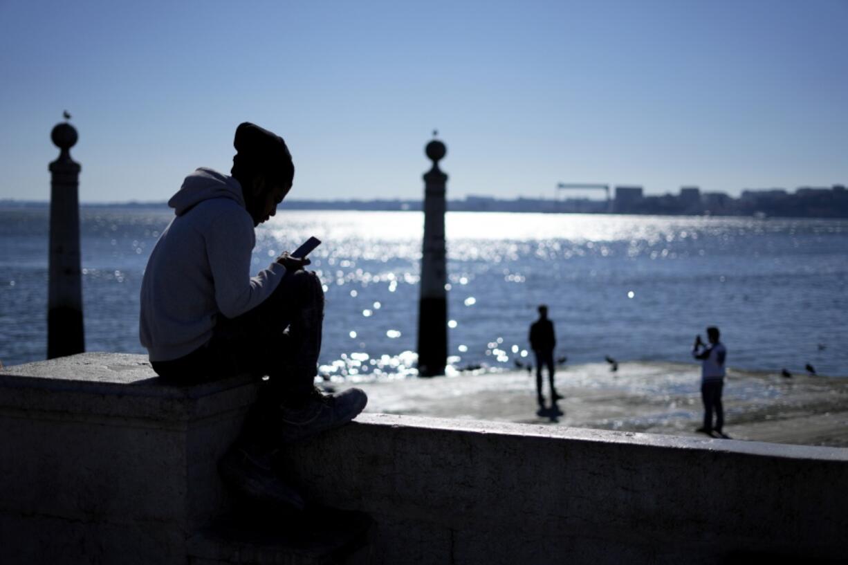 A young man checks his phone by the Tagus river at Lisbon's Comercio square on a sunny winter day, Monday, Jan. 30, 2023.