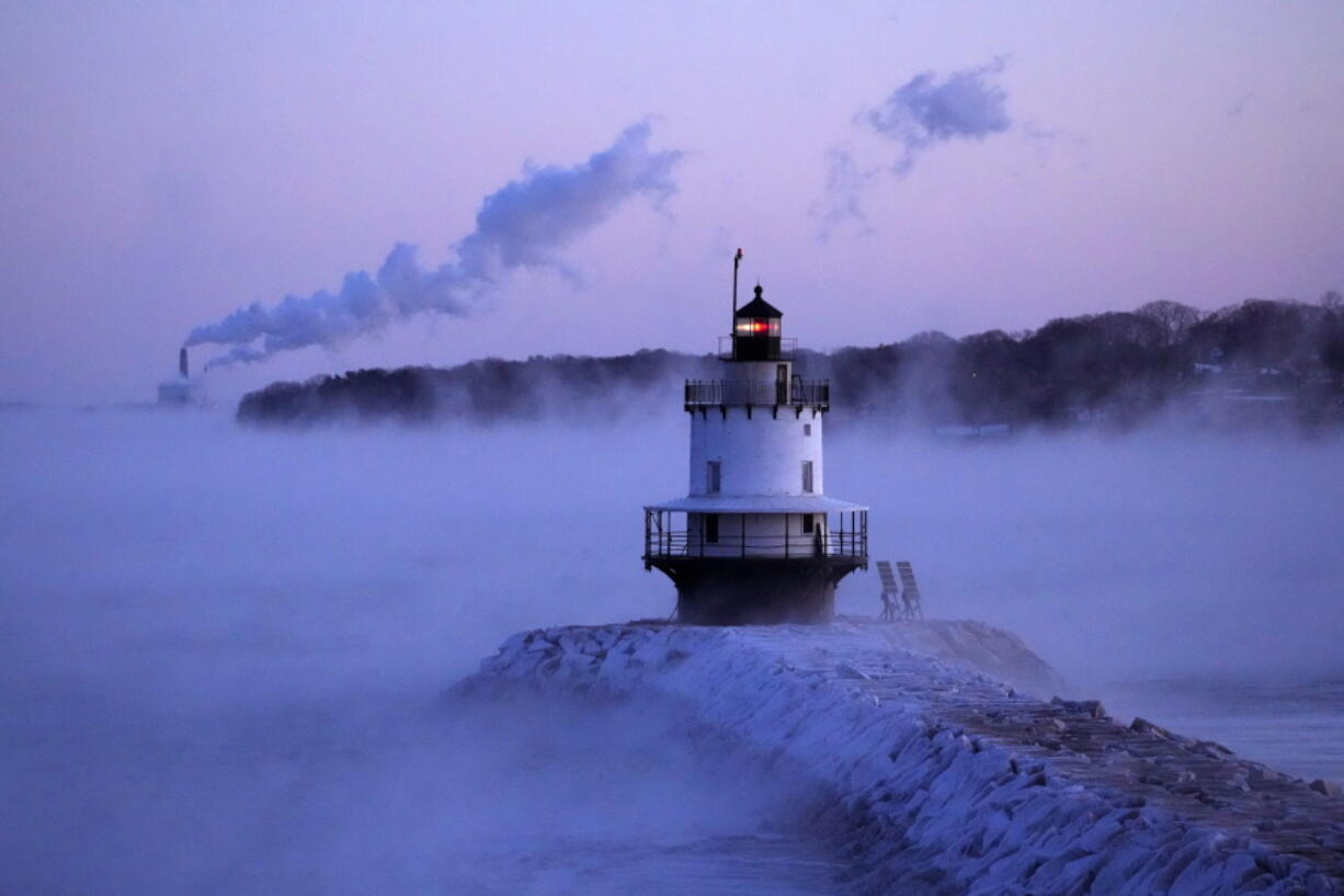 Spring Point Ledge Light is surrounded by arctic sea smoke while emissions from the Wyman Power plant, background, are blown horizontal by the fierce wind, Saturday, Feb. 4, 2023, in South Portland, Maine. The morning temperature was about -10 degrees Fahrenheit. (AP Photo/Robert F.