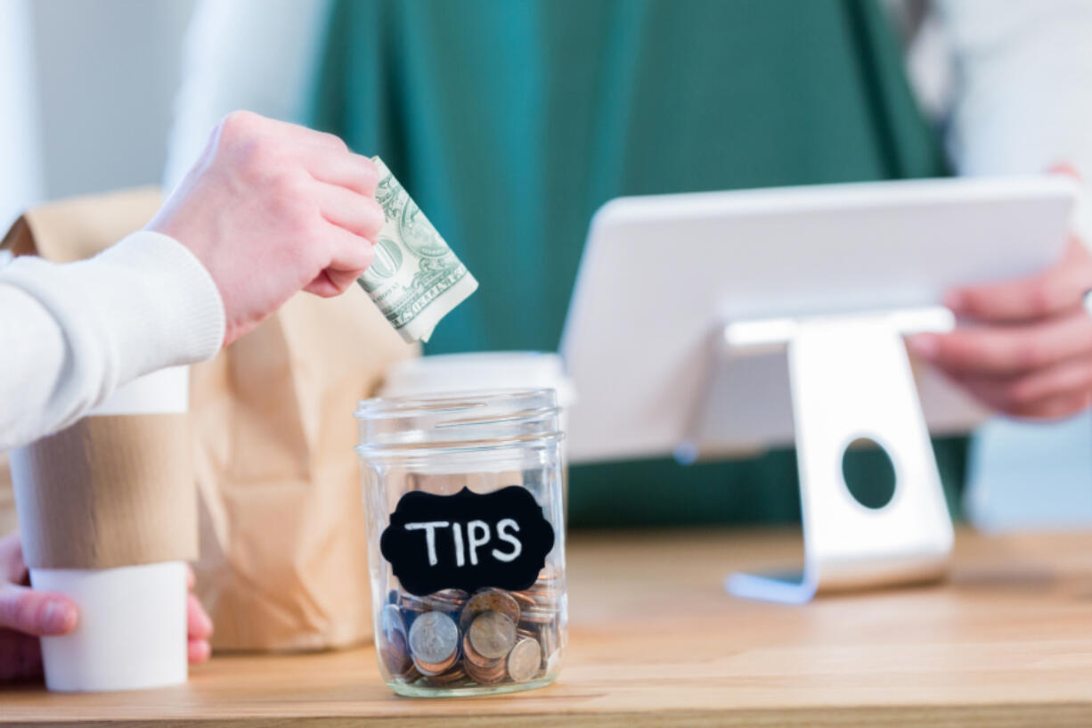 An unrecognizable coffee shop customer stands across the checkout counter from an unrecognizable barista and reaches out to put a paper bill in the tips jar.