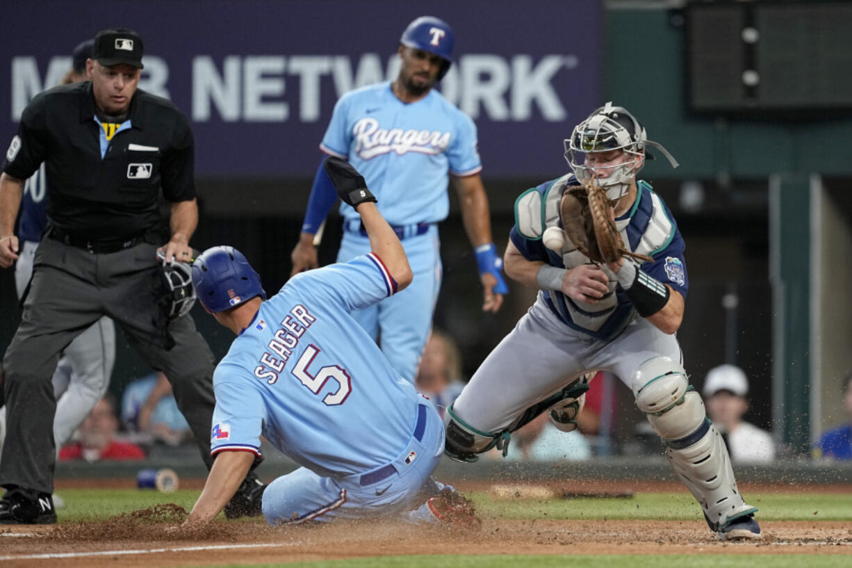 Texas Rangers' Corey Seager (5) scores on a Jonah Heim single as Seattle Mariners catcher Cal Raleigh reaches out for the throw to the plate in the first inning of a baseball game, Sunday, June 4, 2023, in Arlington, Texas. Umpire Andy Fletcher watches the play.