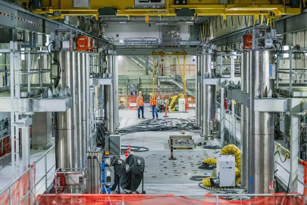 Hanford workers stand near the top of the first melter, flush with the floor in the foreground. The large black hoses of the startup heaters are inserted into the melter.