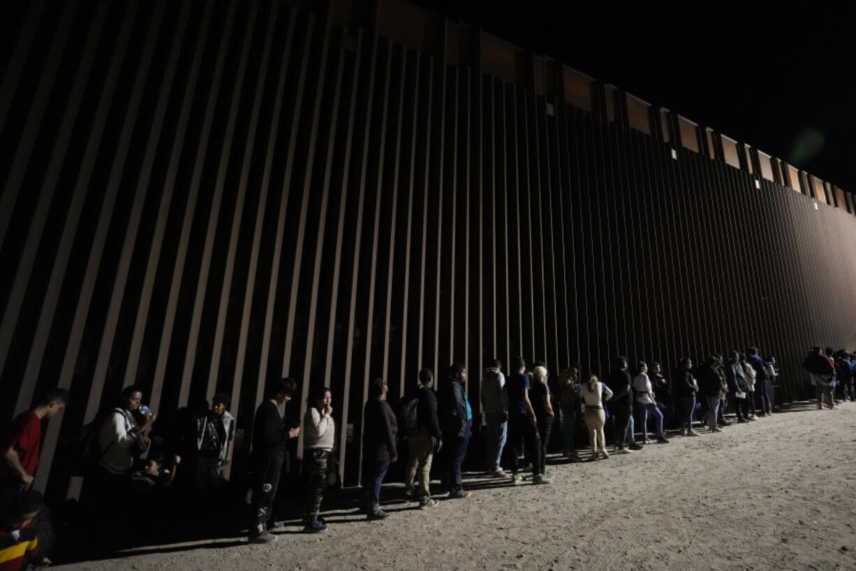 FILE - People line up against a border wall as they wait to apply for asylum after crossing the border from Mexico. Tuesday, July 11, 2023, near Yuma, Ariz. As the number of migrants coming to the U.S.'s southern border is climbing, the Biden administration aims to admit more refugees from Latin America and the Caribbean over the next year. The White House Friday, Sept. 29, released the targets for how many refugees it aims to admit over the next fiscal year starting October 1 and from what regions of the world.