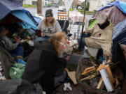 Cheri Rhodes, gray beanie, joins Mike Moises, white glasses, and other members of their street family as a small fire provides a little warmth in the pouring rain at their downtown Vancouver encampment on a recent afternoon.