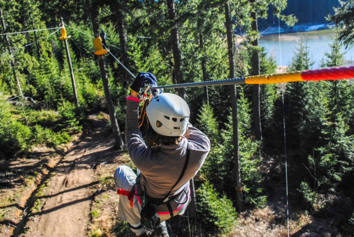 A woman glides down a zip line over a wooded area. The Catamount Zip Tour transports riders from the top of a mountain via a cable suspended hundreds of feet above the trees, a 5,523-foot line, a bit more than a mile.
