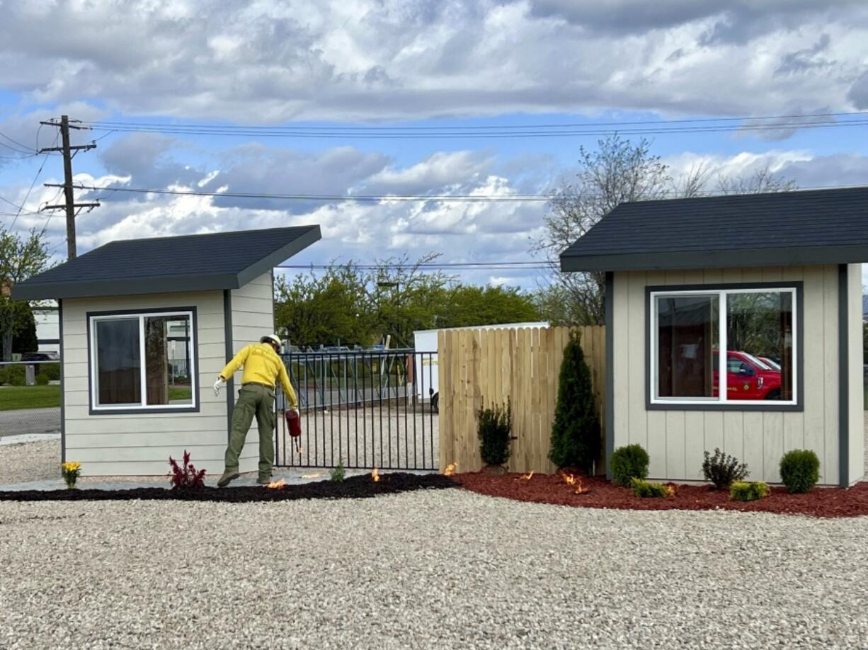 A firefighter uses a drip torch to simulate blowing embers during a burn demonstration for the National Association of Insurance Commissioners Western Zone at the National Interagency Fire Center in Boise, Idaho, Monday, April 29, 2924. The building on the left is designed to mimic a home built using fire-resistant materials with defensible space around the exterior, and the building on the right is designed to mimic a home with traditional landscaping and building materials. The building on the right quickly burned to the ground.