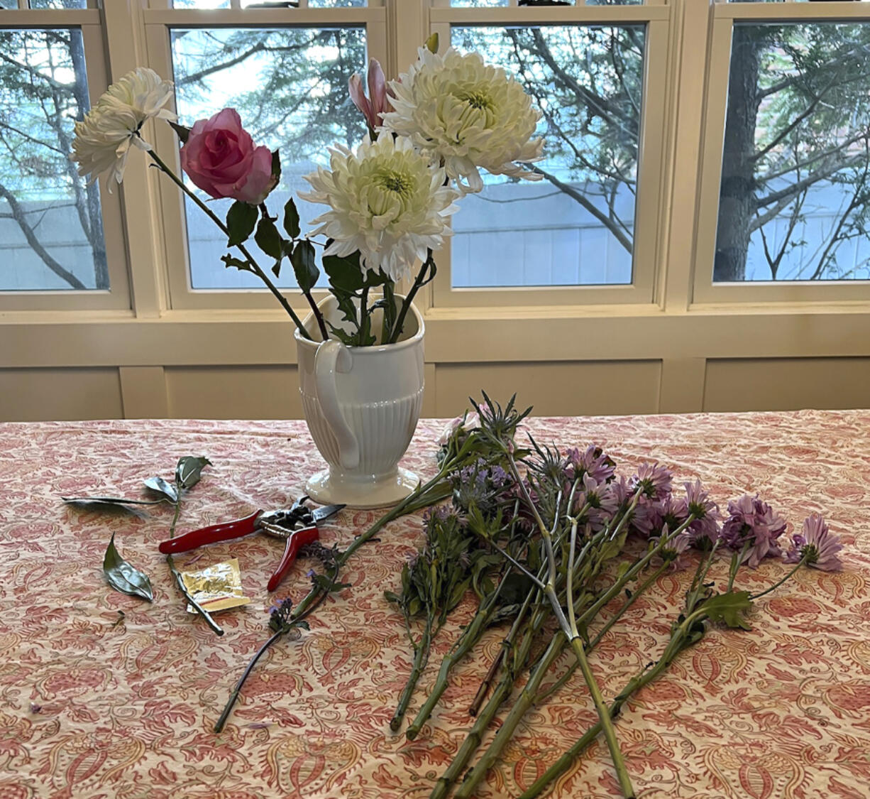 A supermarket bouquet of three white chrysanthemums, a lily stem and a single rose in a decorative pitcher, along with garden pruners and a packet of flower-preserving granules.