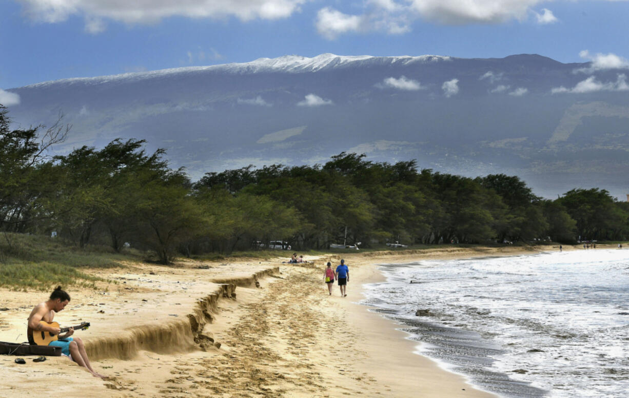 FILE -- Snow-capped Haleakala serves as a backdrop as Paia, Maui&rsquo;s Scott Picton, left, plays a guitar and Saskatchewan&rsquo;s Shannon and Dan Runcie walk on Sugar Beach in South Maui, Hawaii, Feb. 11, 2019. Local officials on the Hawaiian island of Maui on Wednesday, June 5, 2024, voted to oppose a U.S. military proposal to build new telescopes on the summit of Haleakala volcano.