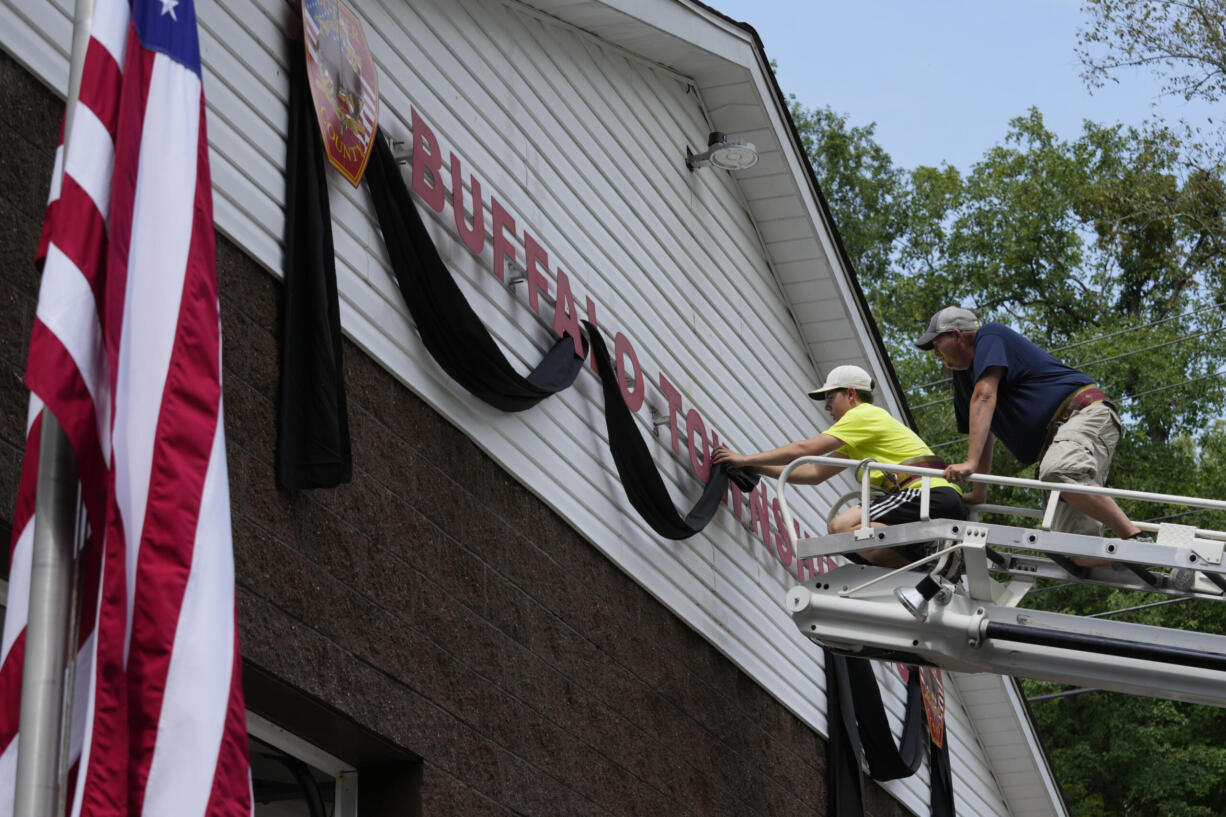 Logan Check, left, junior firefighter, and Randy Reamer, president and rescue captain at the Buffalo Township Fire Company 27, hang bunting on the fire station in memory of fellow firefighter Corey Comperatore, in Buffalo Township, Pa., Sunday, July 14, 2024. Comperatore was killed during a shooting at a campaign rally for Republican presidential candidate former President Donald Trump in Butler, Pa., on Saturday. The flag at the station house flies at half staff at left.