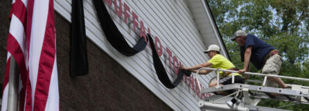 Logan Check, left, junior firefighter, and Randy Reamer, president and rescue captain at the Buffalo Township Fire Company 27, hang bunting on the fire station in memory of fellow firefighter Corey Comperatore, in Buffalo Township, Pa., Sunday, July 14, 2024. Comperatore was killed during a shooting at a campaign rally for Republican presidential candidate former President Donald Trump in Butler, Pa., on Saturday. The flag at the station house flies at half staff at left.