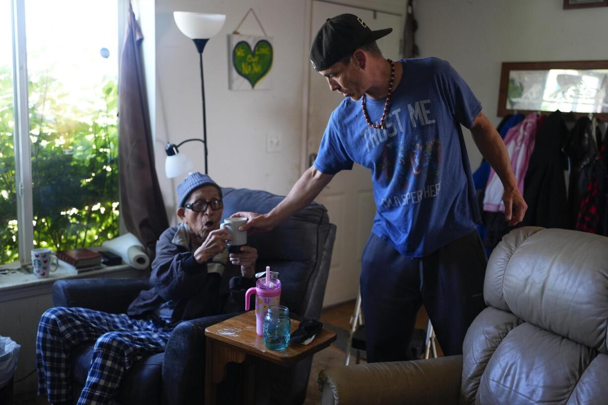 FILE - Sonny Curley hands a cup of coffee to his father Harold Curley at their home near the ocean Wednesday, May 22, 2024, on the Quinault reservation in Taholah, Wash. The home has a rotting deck and black mold inside, and the family has had to evacuate several times due to flooding.