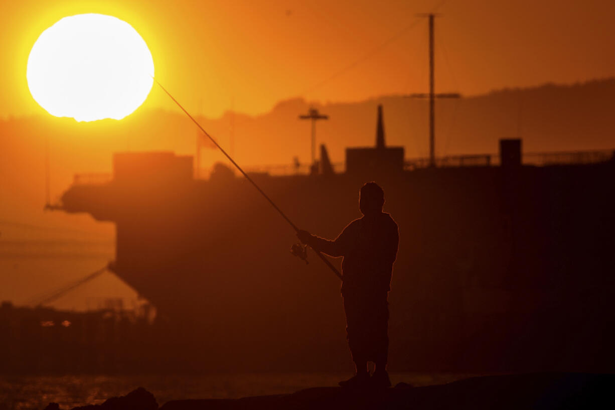 A man fishes off a jetty in Alameda, Calif., as the sun sets over the San Francisco Bay on Monday, July 1, 2024. An extended heatwave predicted to blanket Northern California has resulted in red flag fire warnings and the possibility of power shutoffs beginning Tuesday.