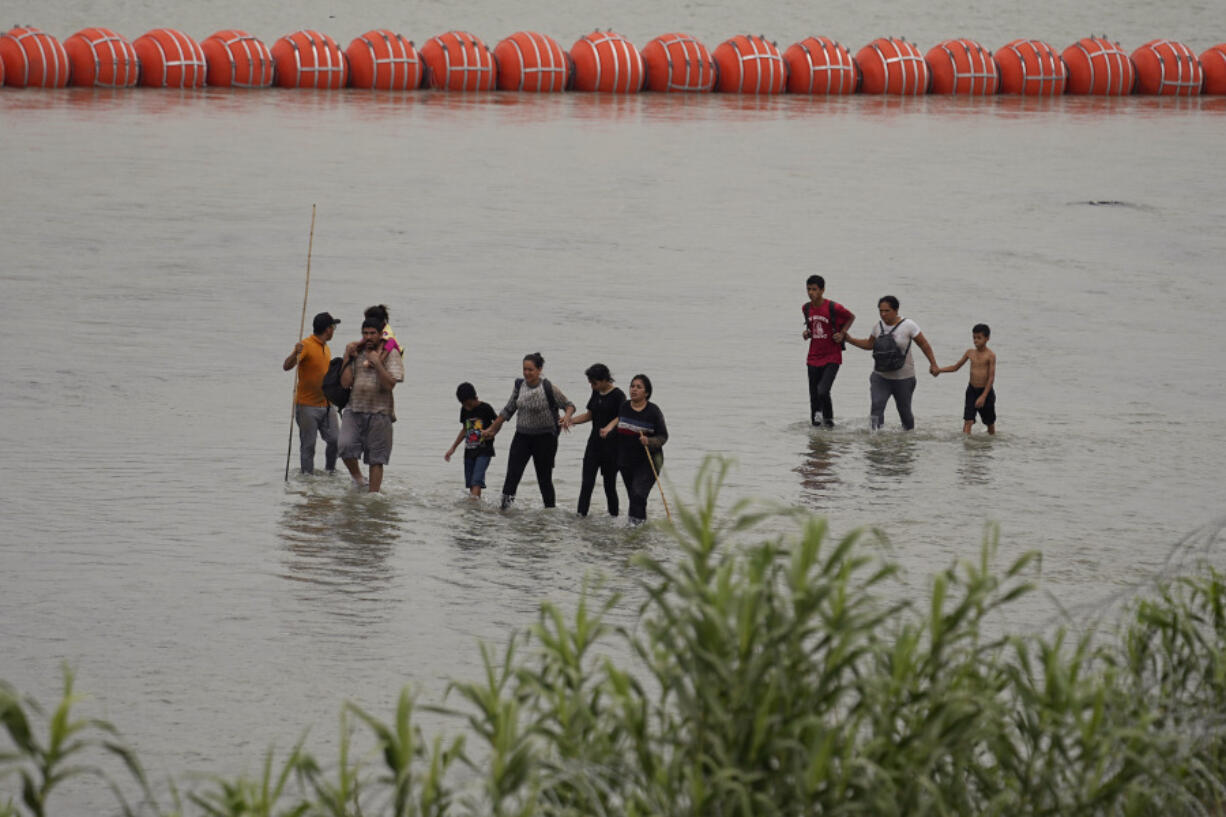 FILE - Migrants who crossed the Rio Grande from Mexico walk past large buoys being deployed as a border barrier on the river in Eagle Pass, Texas, July 12, 2023. The floating barrier is being deployed in an effort to block migrants from entering Texas from Mexico.