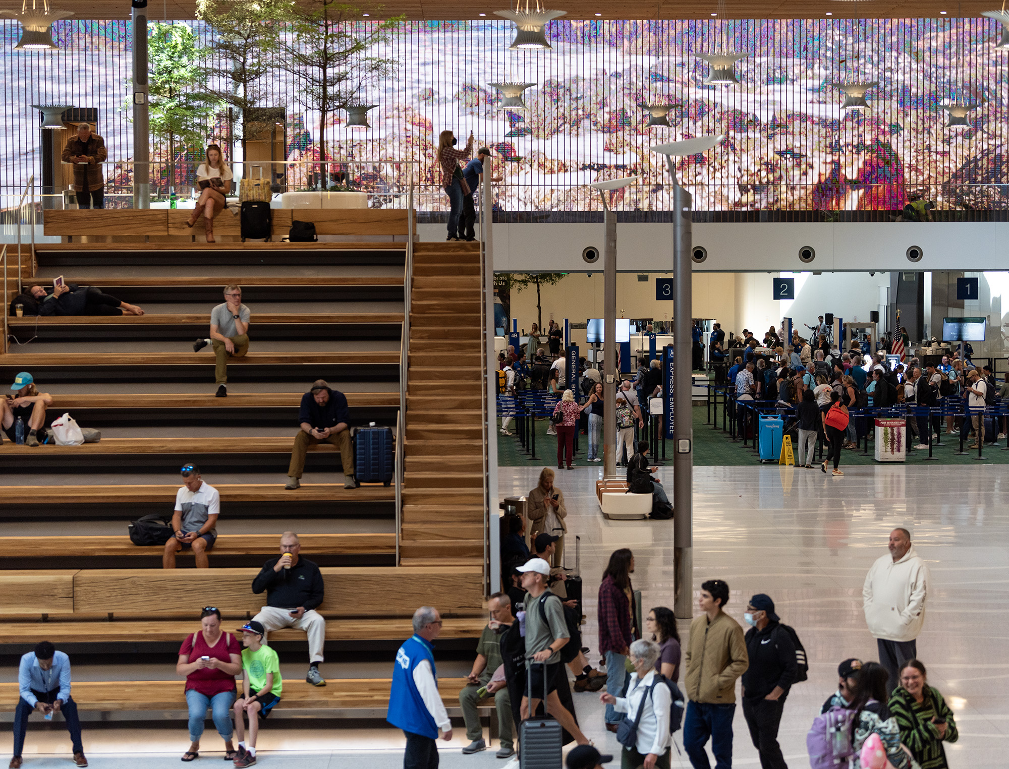 Travelers lounge on stadium seating and move through security underneath a giant video board Wednesday, Aug. 14, 2024, at Portland International Airport. Opening the new main terminal to the public marked the end of phase 1 of the PDX Next project, with phase 2 and the completion of all construction scheduled to wrap up in Spring 2026.