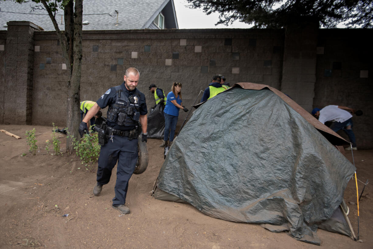 Vancouver police Cpl. Sean Metevia, left, joins cleanup crews and members of the city of Vancouver&rsquo;s Homelessness Assistance and Resource Team on July 31 as they remove solid waste along the West Mill Plain Boulevard sound wall, with a house just beyond. Residents on the other side of the sound wall want the camp cleared.