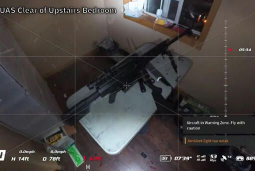 A screenshot from drone footage shows a rifle set up on a table in an upstairs bedroom.