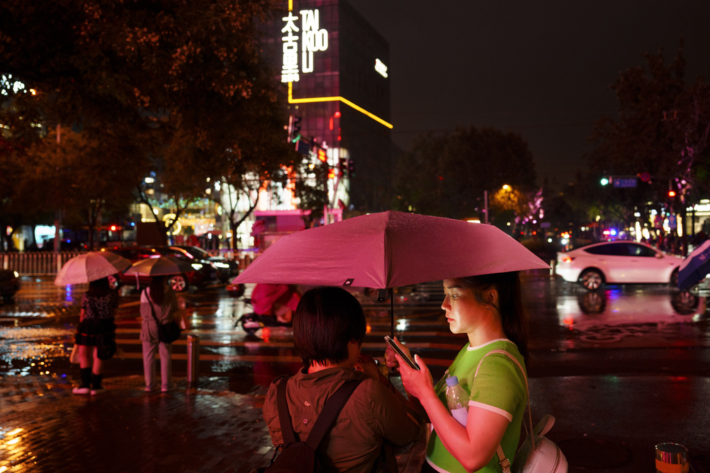 FILE - People look at their phones while waiting to cross an intersection in the rain at the Taikoo Li Sanlitun shopping center in Beijing, July, 30, 2024.