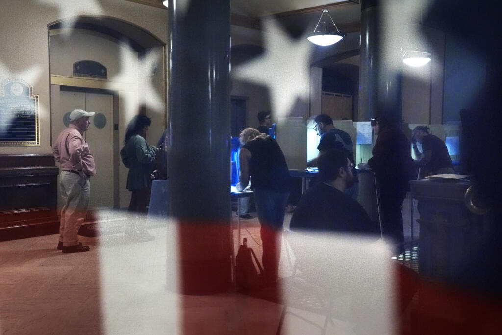 Voters are reflected in a window near an American flag as they mark their ballots during early voting in the general election, Friday, Nov. 1, 2024, at City Hall in Providence, R.I.